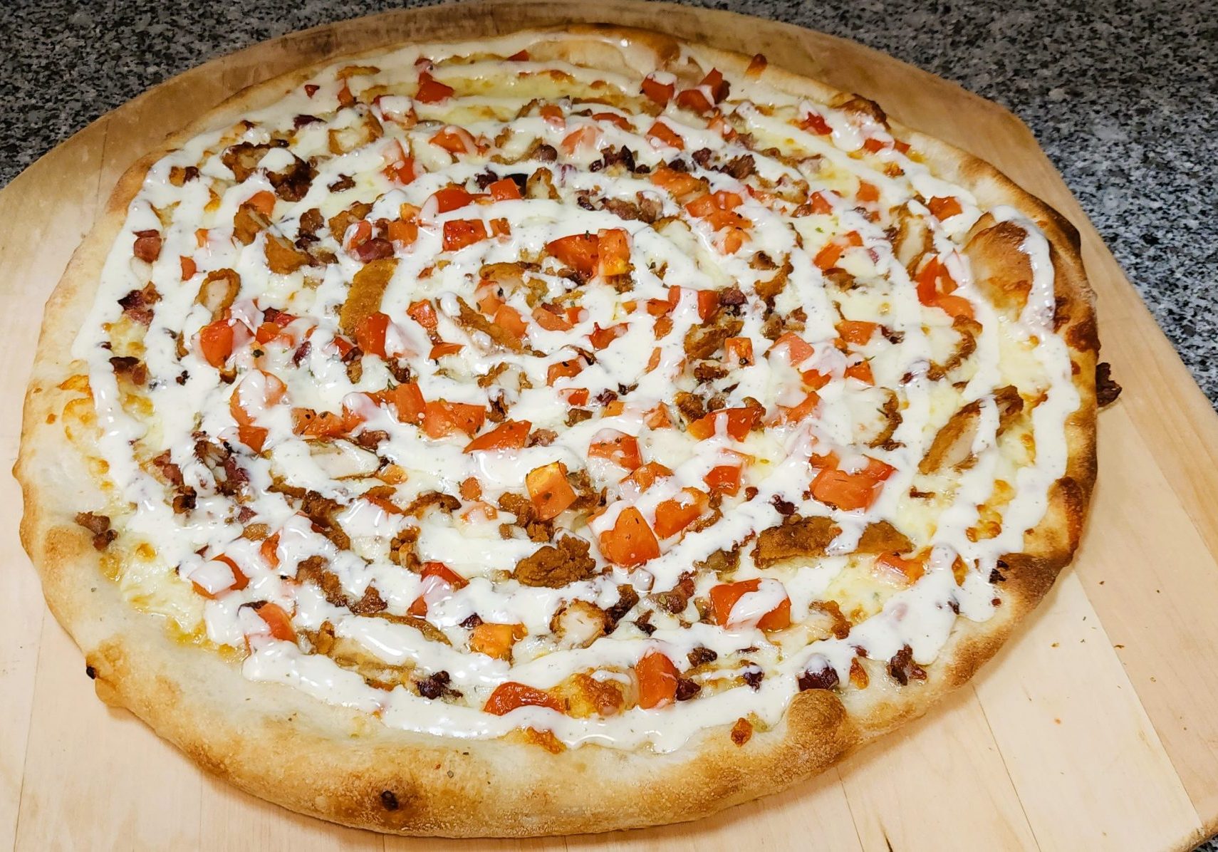 A freshly baked pizza topped with a generous amount of diced tomatoes, meat, and a swirl of white sauce on a baking board.