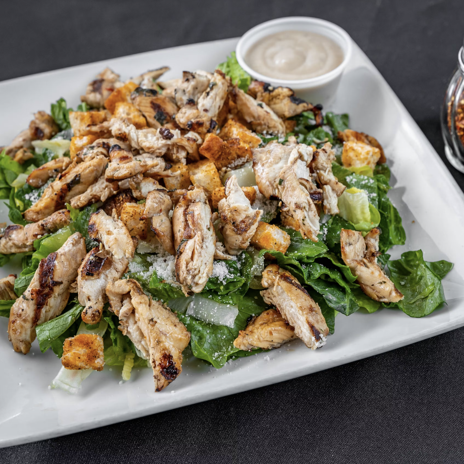 A plate of chicken Caesar salad with grilled chicken, croutons, Parmesan cheese, and romaine lettuce, served with dressing on the side. Salt and pepper shakers are placed nearby.