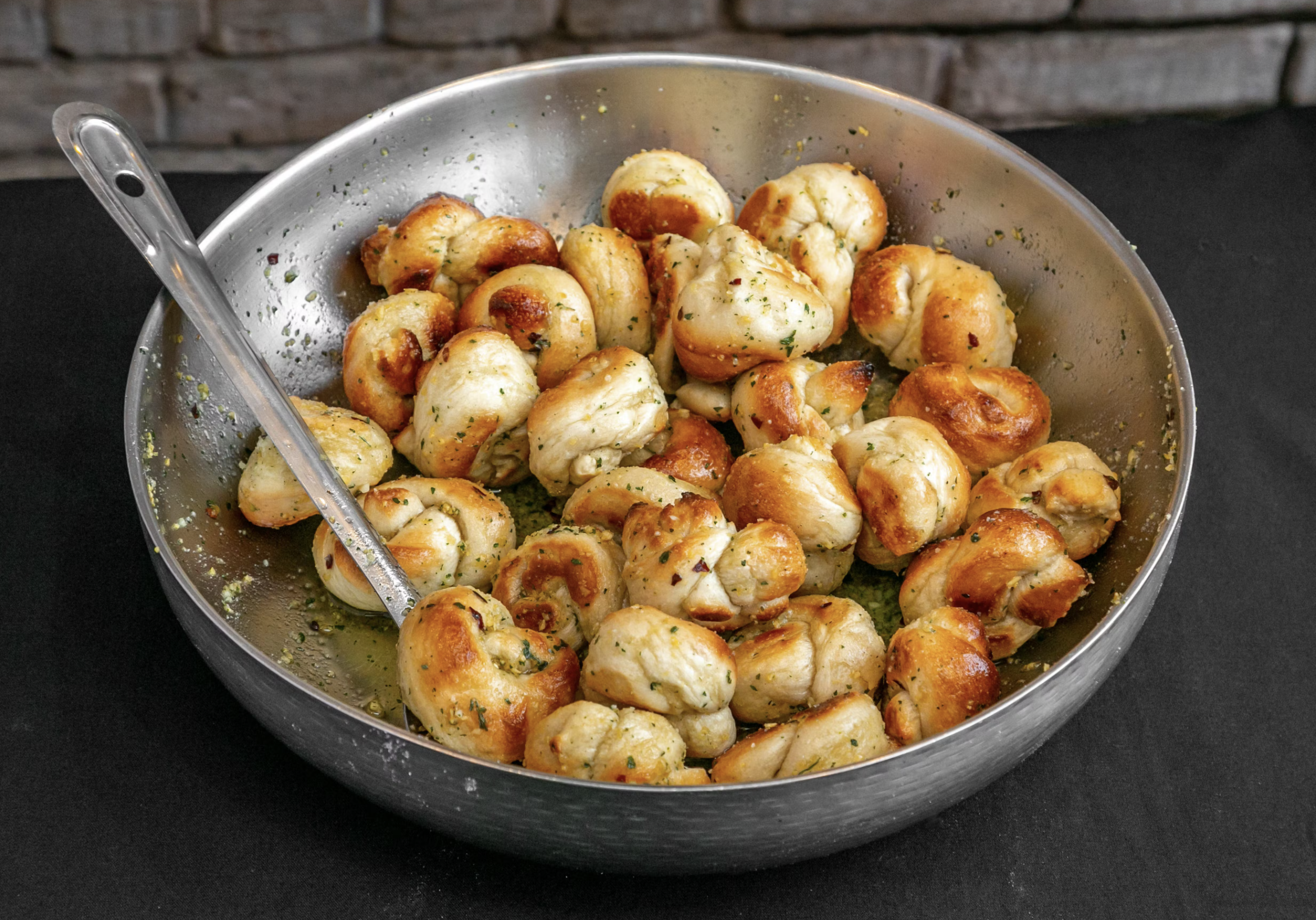 A silver bowl containing seasoned garlic knots with a serving spoon placed inside.